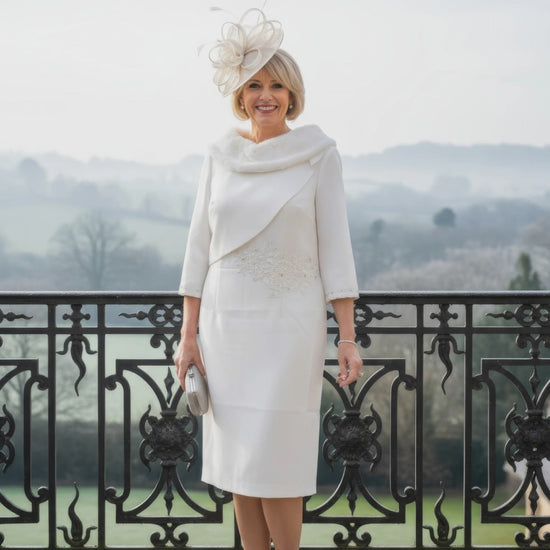Woman in a white fur bridal dress with a matching hat standing on a balcony with a scenic background.