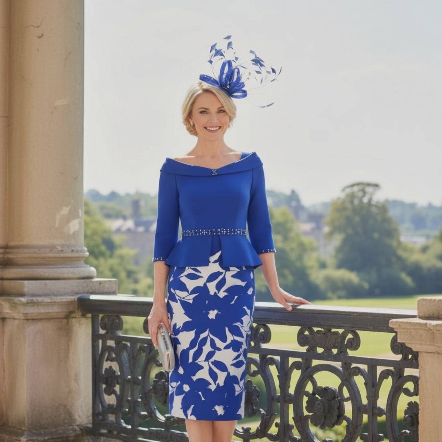 Woman in a Gino Cerrutti cobalt blue  mother of the bride dress with a matching cobalt blue fascinator standing on a balcony with a scenic background. Signature Dress.