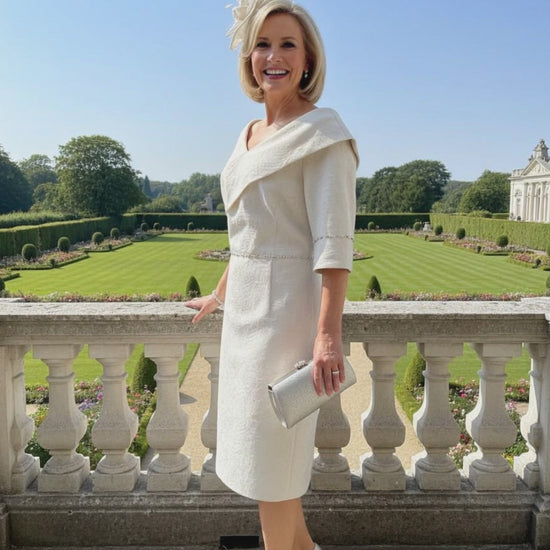 Woman in a lizabella gold sheath mother of the bride dress with a floral headpiece standing outdoors with a scenic background
