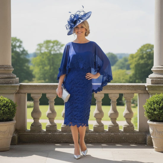 Woman in a veni infantino blue mother of the bride dress and poncho wearing a matching fascinator hat standing on a balcony with greenery in the background. Signature Dress.