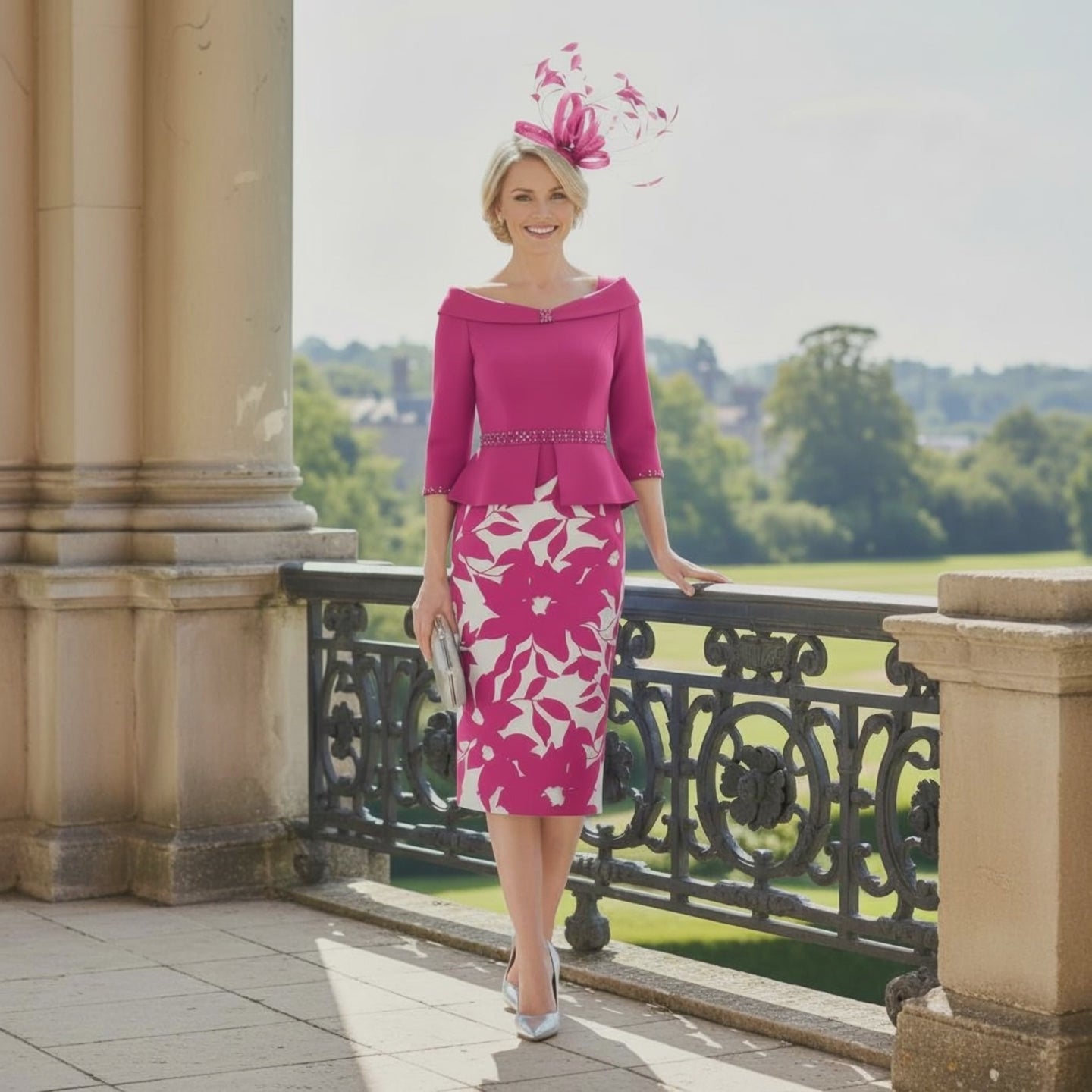 Woman in a Gino Cerrutti pink mother of the bride outfit with a floral hat standing on a balcony with a scenic background. Signature Dress.