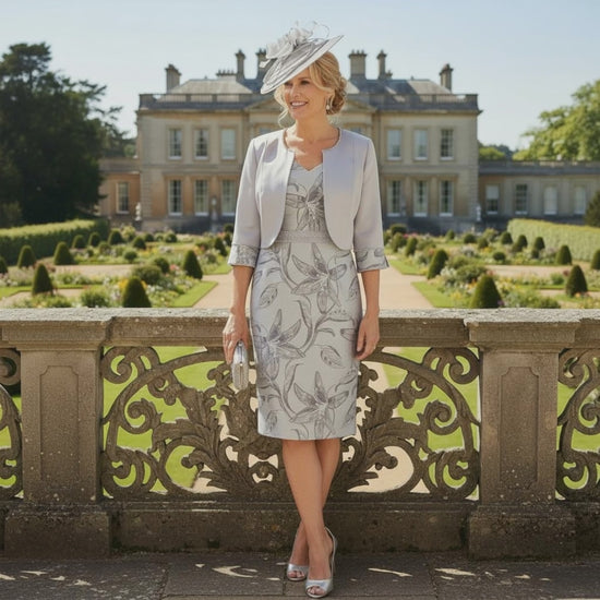Woman in a floral Lizabella Silver 2 piece mother of the bride dress and matching jacket with a silver bespoke fascinator by Signature Dress standing in front of a large building with gardens. Signature Dress.