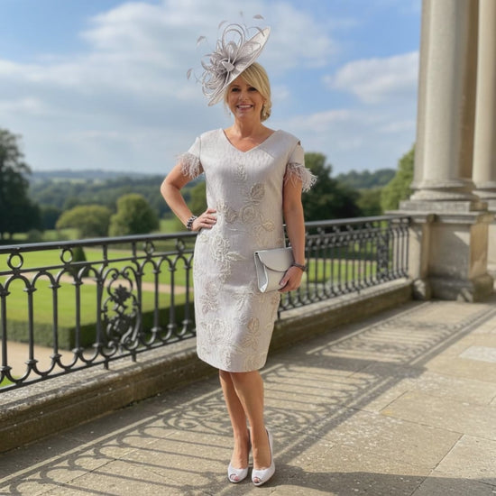 Woman in Lizabella a light lilac mother of the bride 2 piece outfit with a matching hat standing on a balcony with a scenic background. Signature Dress.
