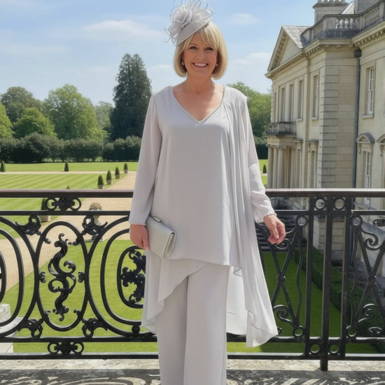 Woman in a silver mother of the bride trouser suit outfit make by veromia, standing in front of a large building with a garden.