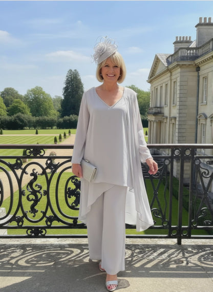 Woman in a silver mother of the bride trouser suit outfit make by veromia, standing in front of a large building with a garden.