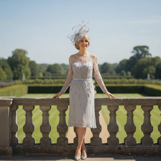 Woman in a Veni Infantino Silver gatsby mother of the bride dress and matching fascinator hat standing on a stone railing with a scenic background.  Signature Dress.