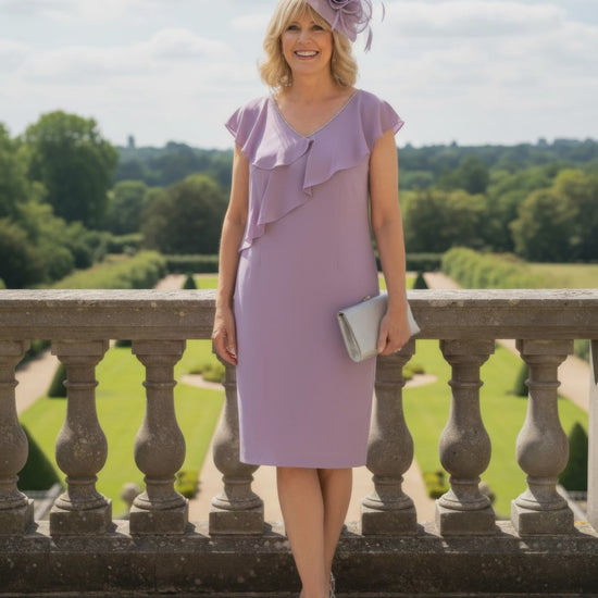 Woman in a lilac mother of the bride dress with ruffled details and a matching hat standing on a stone balcony with a garden view.