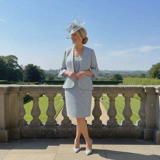 Woman in a plus size Veromia light pastel blue mother of the bride 2 piece suit with a matching fascinator hat standing on a stone ready to attend a wedding. 