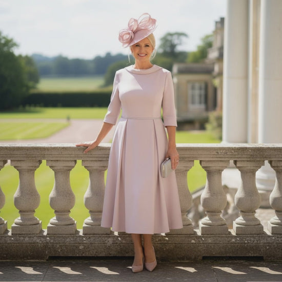 Woman in a Veromia light pink mother of the bride dress and hat standing on a balcony, the dress is midi length with 3/4 sleeves.