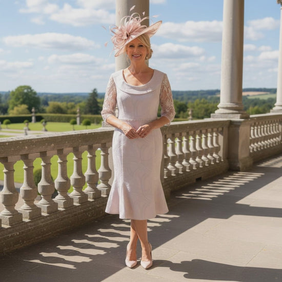 Woman in a Lizabella blush pink mother of the bride dress and blush pink fascinator hat standing on a balcony with a scenic view. Signature Dress