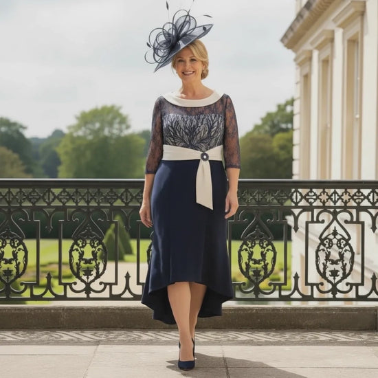 navy Couture Club dress with lace bodice, 3/4 sleeves and high‑low hemline, worn with a navy fascinator on a balcony. Signature Dress