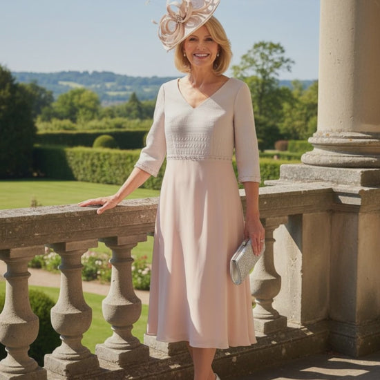 Woman in a light pink Mother of the bride dress by Lizabella with a decorative hat standing on a balcony with a scenic background.