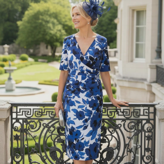 Woman in a Ispirato blue mother of the bride dress standing on a balcony with greenery and a building in the background. Signature Dress.