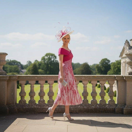 Woman in a pink and white john charles dress with a matching hat standing on a balcony with greenery in the background