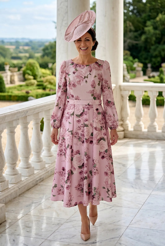 Woman in a veromia pink floral mother of the bride dress and matching pink fascinator standing on a balcony with a garden view