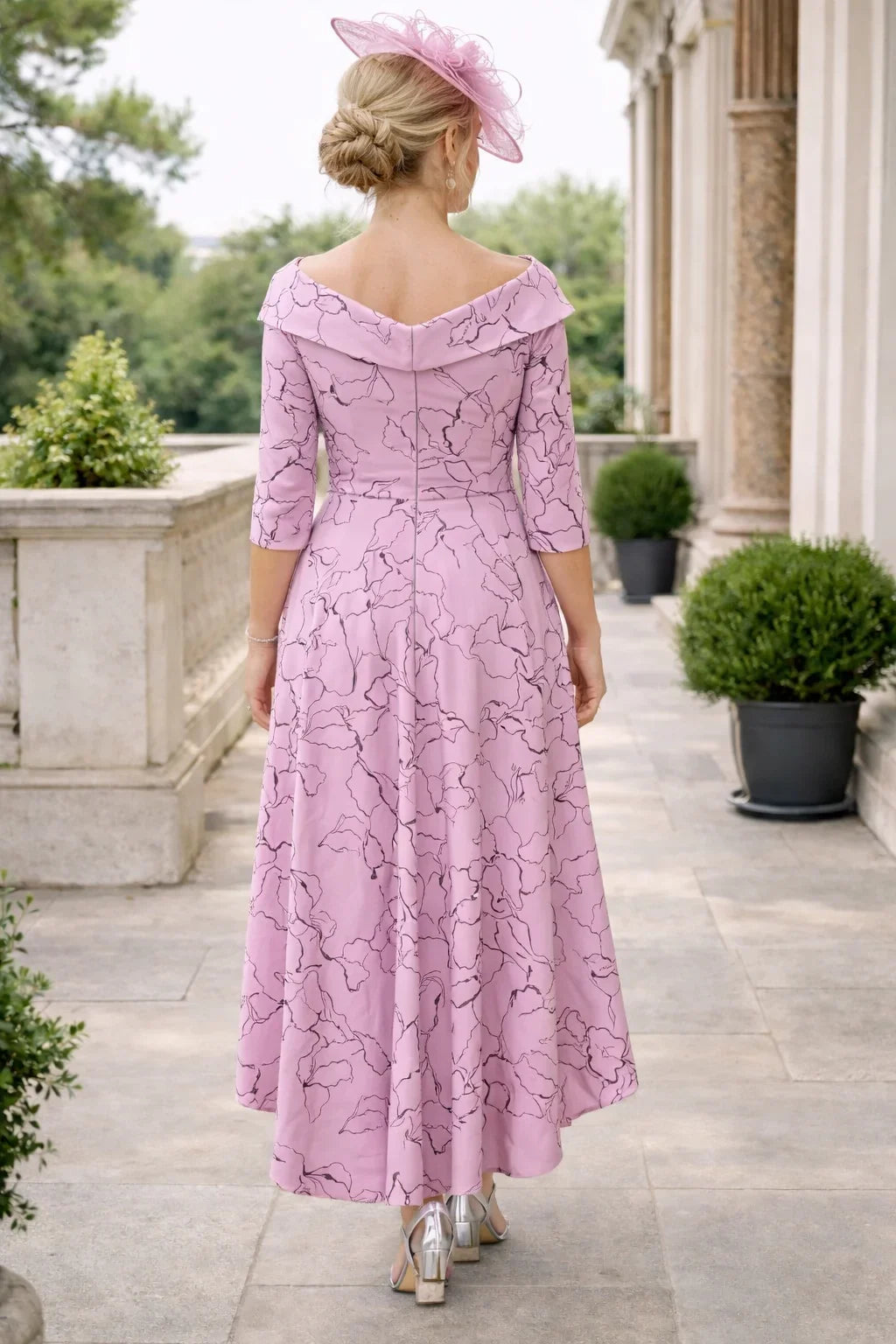 back view of a Woman in a Veromia pink high low dress with a matching pink fascinator hat standing outdoors.