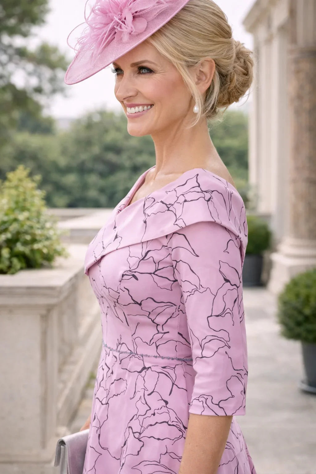 Side close up view of a woman in a Veromia pink high low dress with a matching pink fascinator hat standing outdoors.
